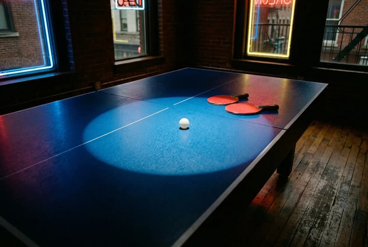 Bright blue ping-pong table under a spotlight in a dark club interior, two paddles and a white ball on the surface, neon strips glowing along the walls behind.