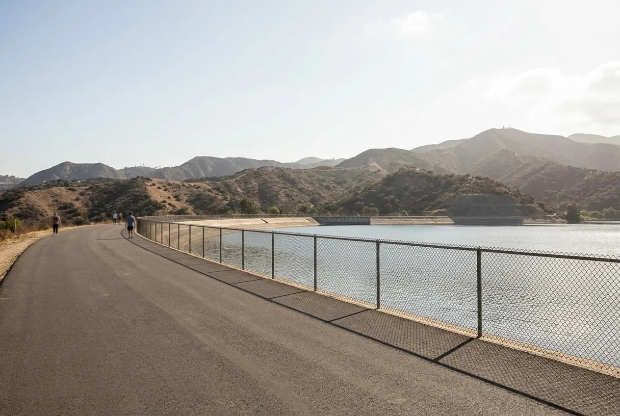 A paved path curving alongside a large urban reservoir with fencing and hills beyond