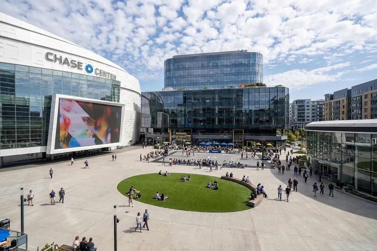 Wide view of Thrive City's open plaza at Chase Center with large outdoor video screen, lawn seating area, and surrounding Mission Bay buildings under clear sky