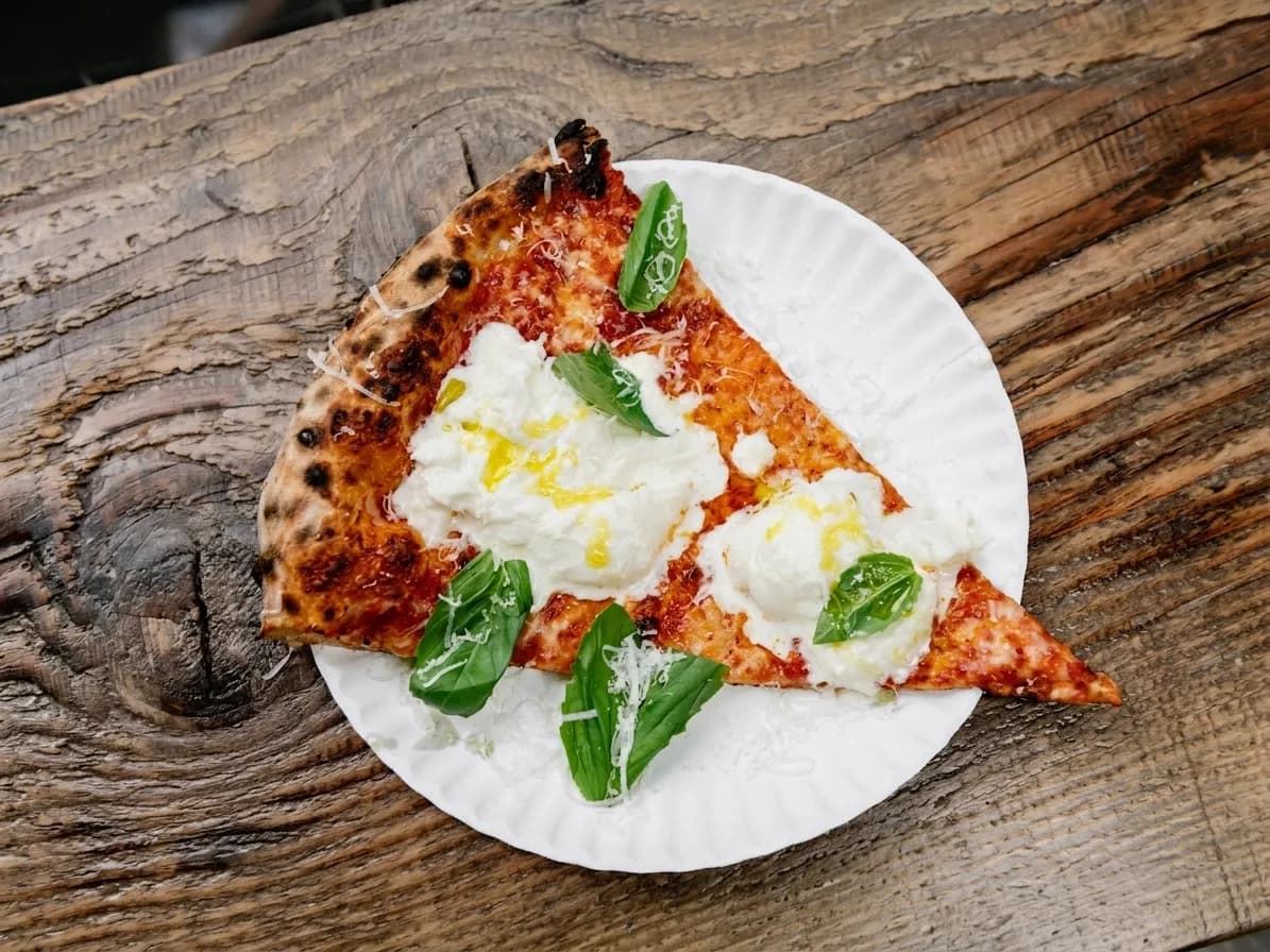 Burrata-topped pizza slice with fresh basil on a paper plate on a wooden counter