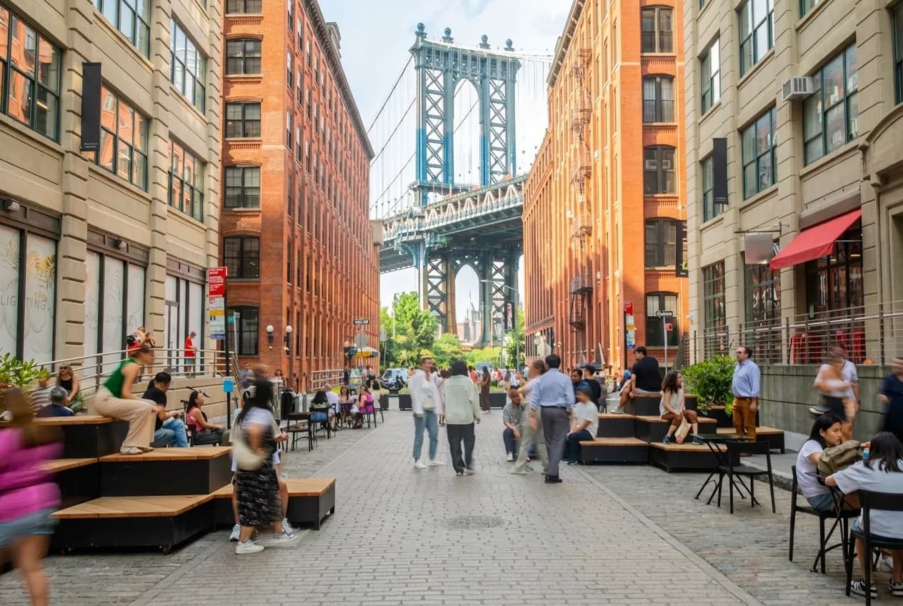 Wide view of a cobblestone Dumbo street with the Manhattan Bridge framed between brick warehouses and people at outdoor tables