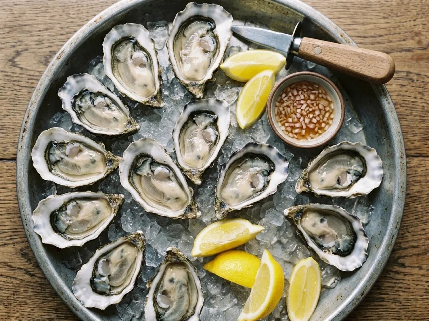 A dozen oysters on crushed ice on a metal tray with lemon wedges and a small dish of mignonette, seen from above