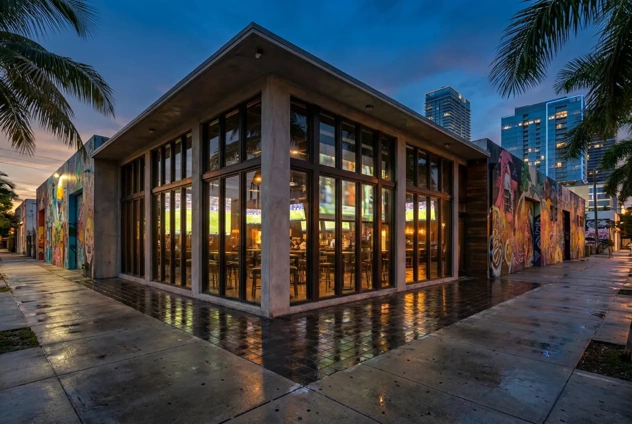 Wynwood warehouse-style sports bar exterior with mural-covered walls and large windows glowing under late-afternoon Miami sunlight, palm shadows on concrete.