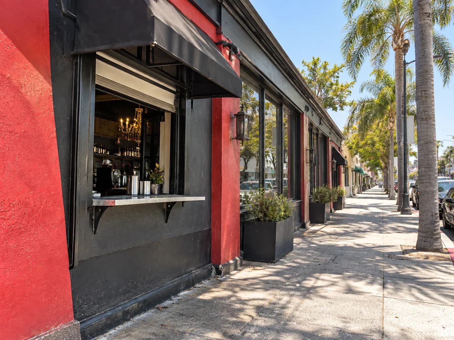 Walk-up service window set into the side wall of a long low restaurant building on a wide sunny avenue, with a small counter ledge under the window and an awning above