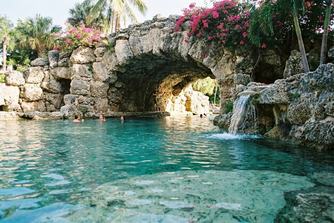 A coral-rock swimming pool in Coral Gables on a bright morning, pale limestone walls and a cave arch behind clear blue-green water with a small cascade