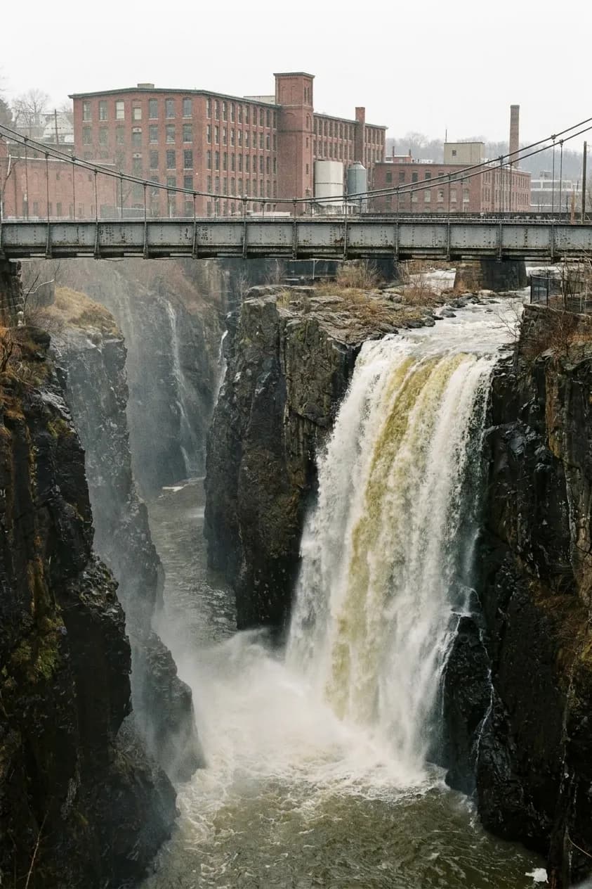 A broad waterfall dropping seventy feet into a basalt gorge with a steel footbridge crossing high above and red brick mill buildings beyond the far rim