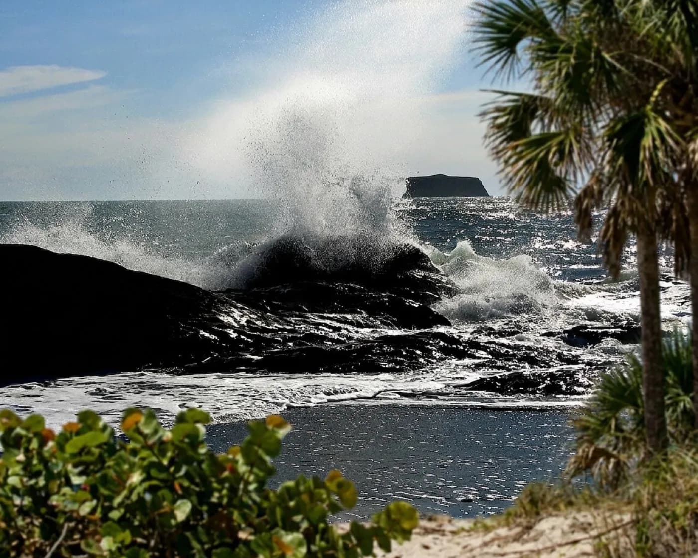 A limestone shoreline with seawater bursting upward through a hole in the rock, spray catching the light against a blue sky