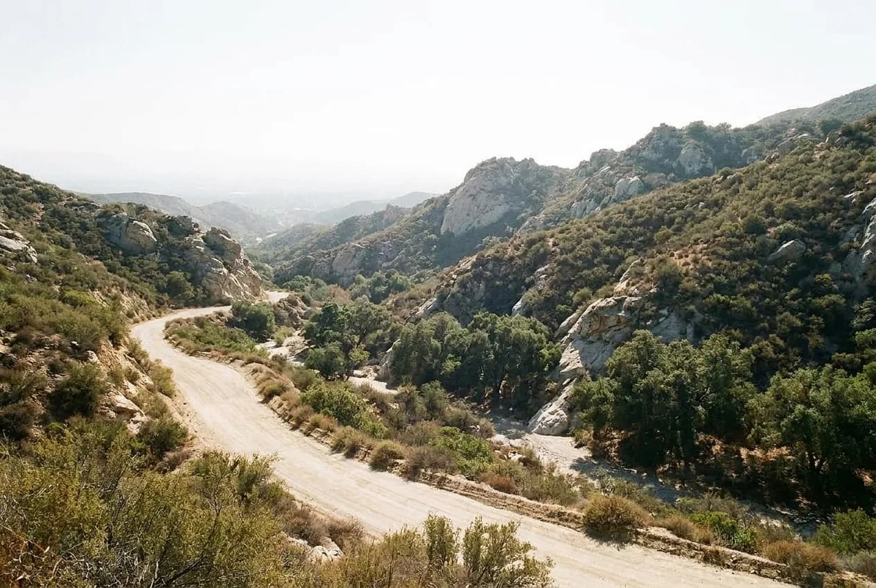 A dirt fire road curving between pale rock outcrops and dry olive-green scrub in a Southern California canyon