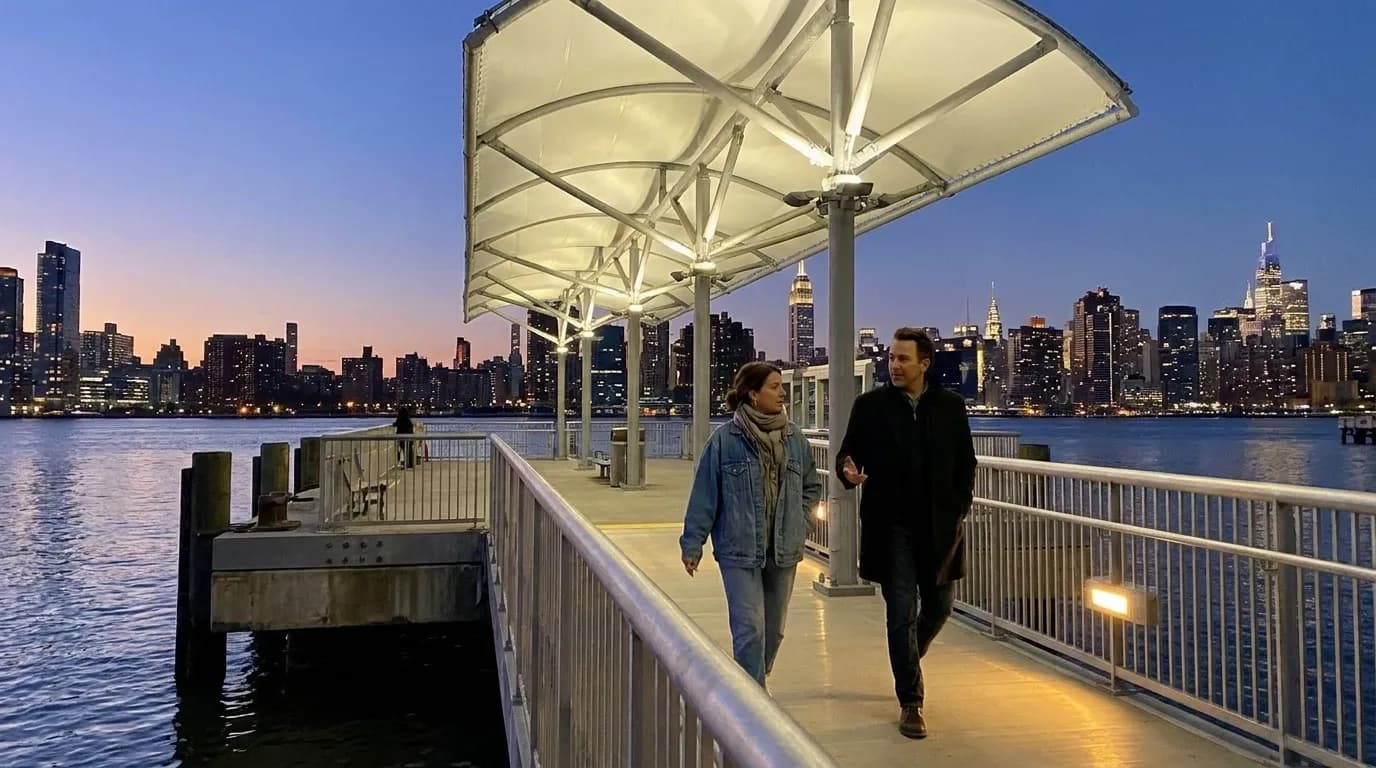 Pedestrians walking along Williamsburg waterfront promenade with East River and city lights in background