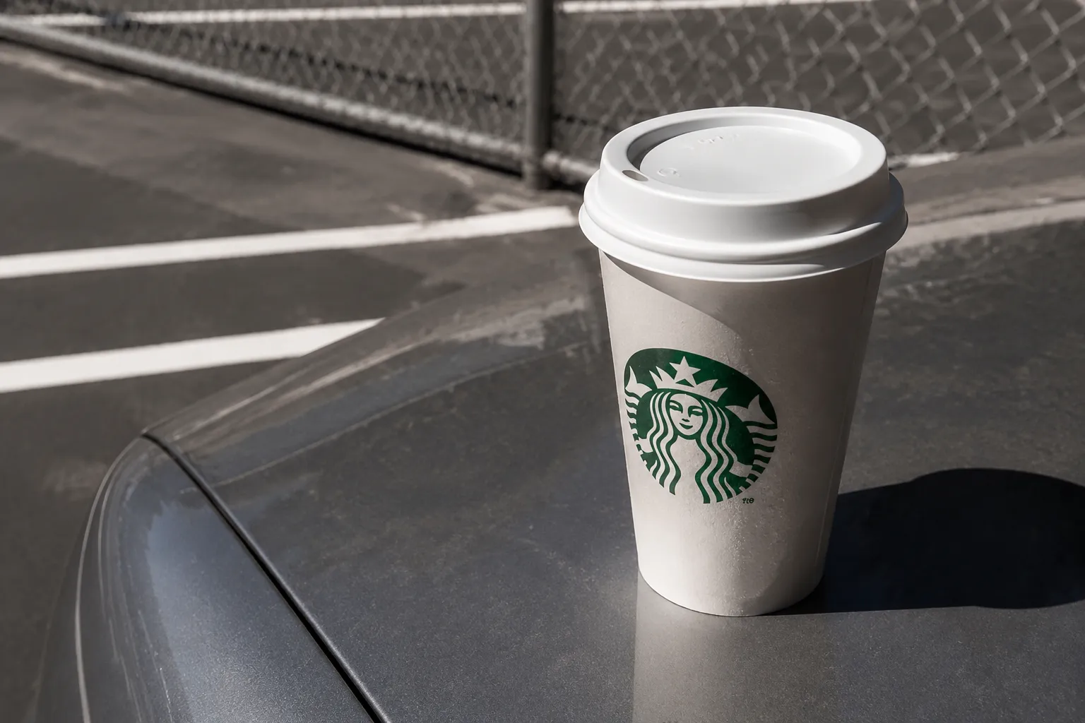 Starbucks to-go cup on a car hood in a Los Angeles parking lot with asphalt and chain-link fence behind