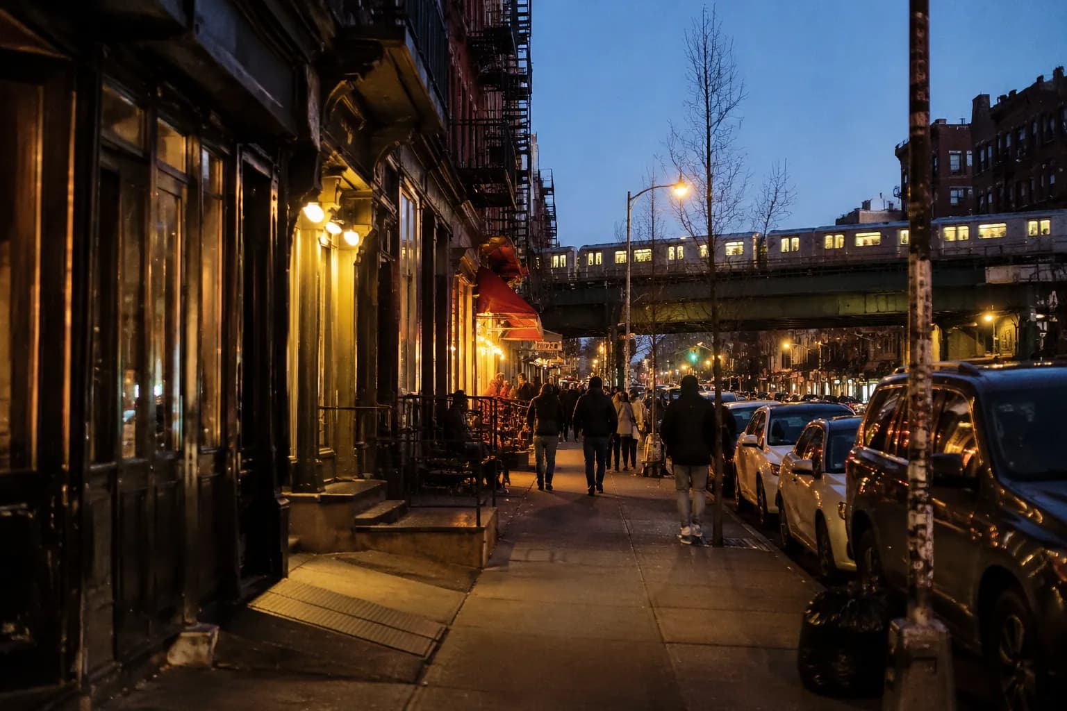 Bedford Avenue in Williamsburg at dusk, with pedestrians and lit storefronts under the elevated L train tracks.