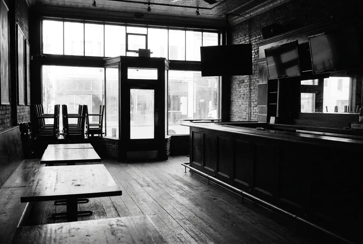A pub interior before opening with chairs still stacked in one corner, screens dark above the bar and pale early light coming through the front glass
