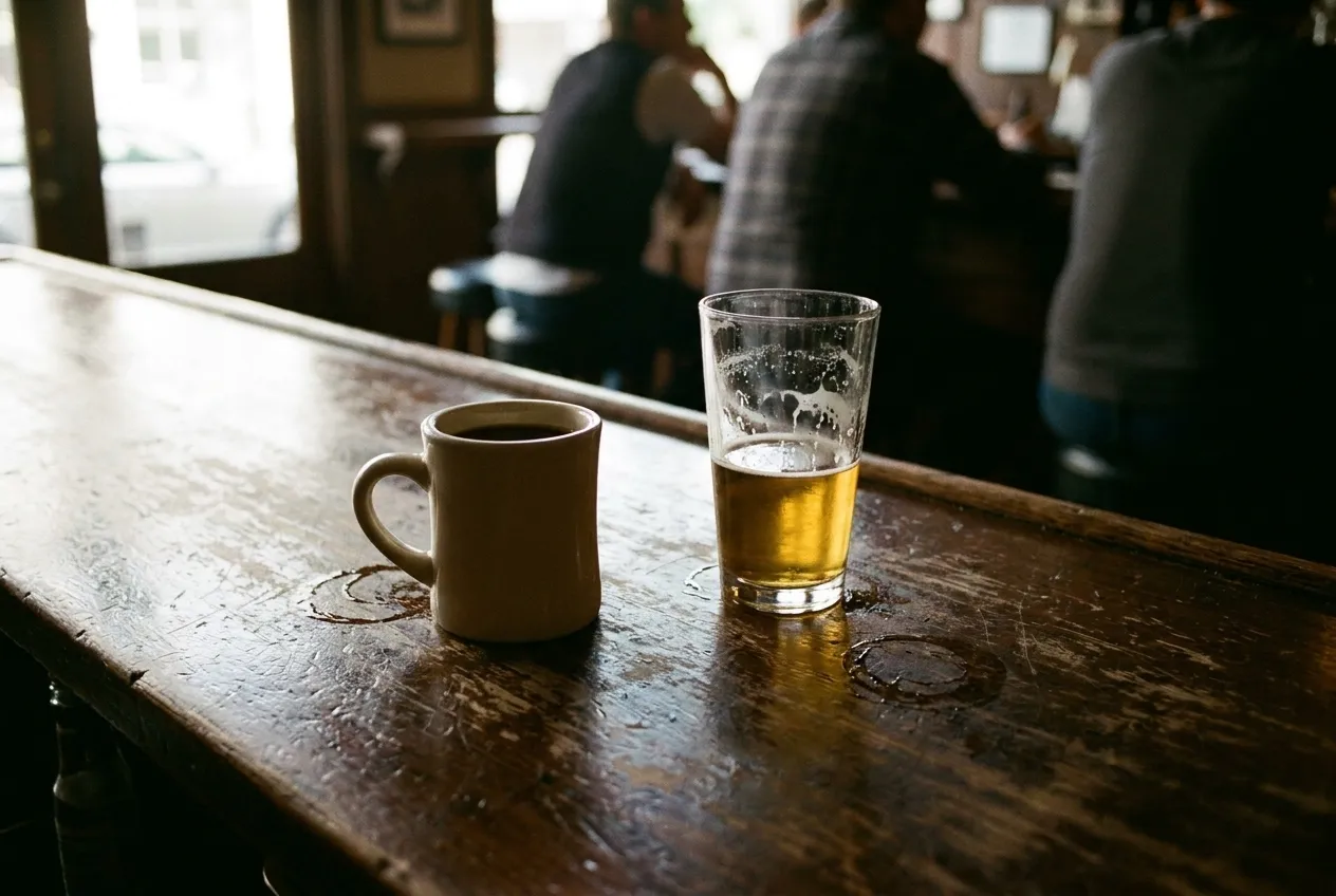 A mug of coffee beside a half-finished pint glass on a worn wooden bar, condensation rings on the surface