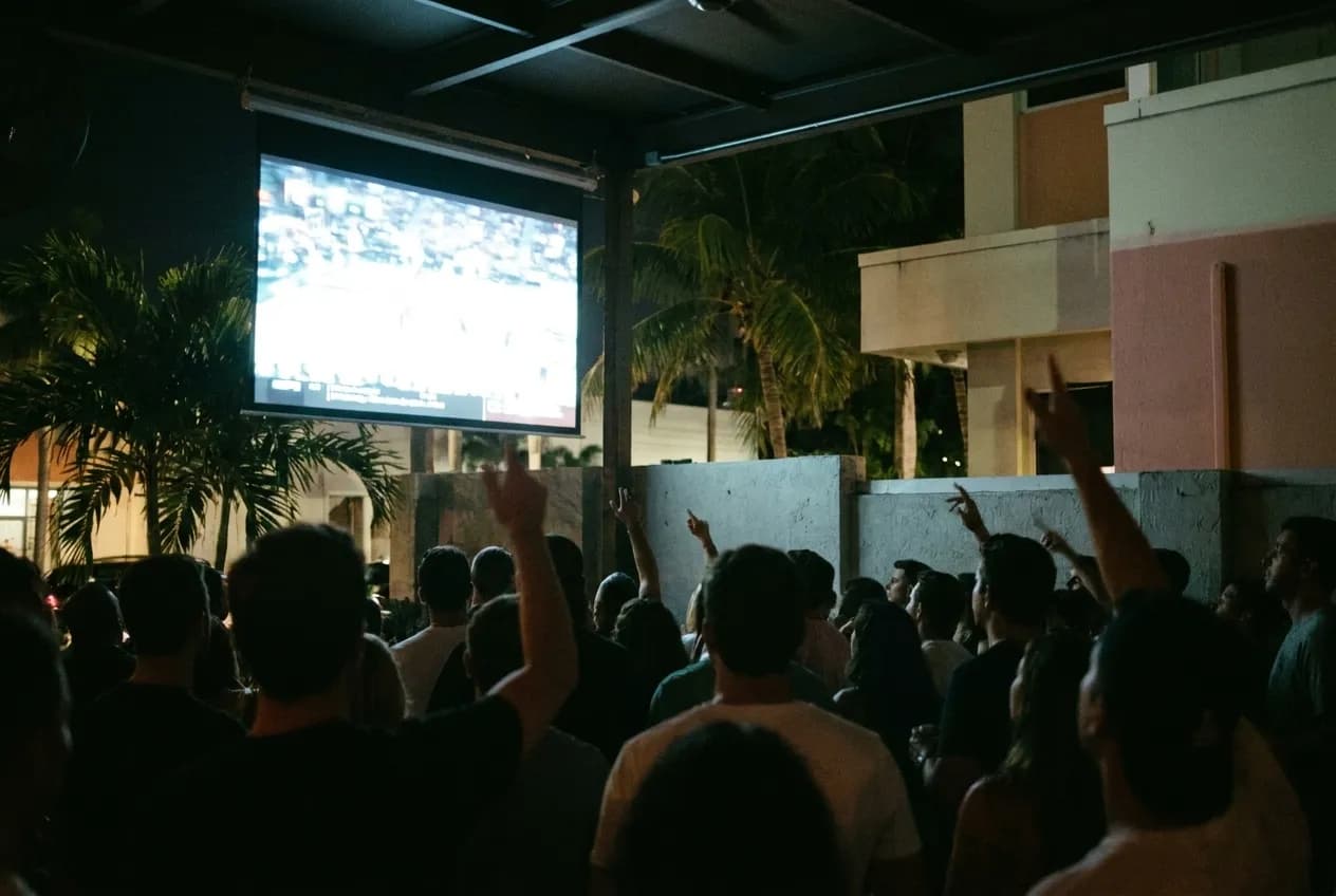 A crowd seen from behind facing a large bright screen in a dim room, silhouetted heads and raised arms across the foreground