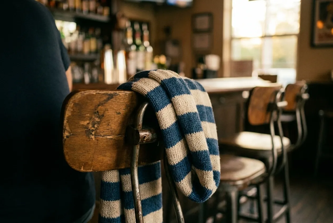 A knitted club scarf in two colours draped over the back of a bar stool, the fabric texture close and the room soft behind