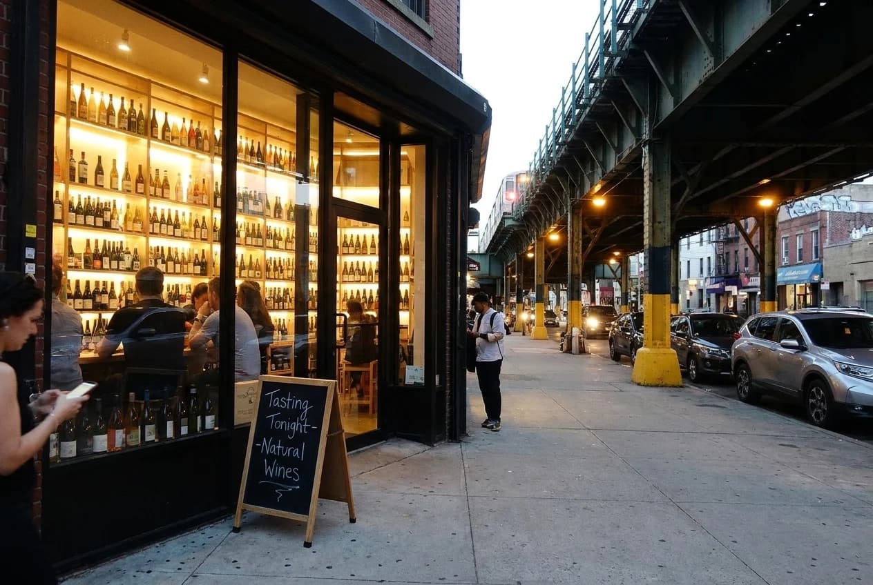 Lit wine shop storefront in Williamsburg at dusk with bottle shelves visible through large windows