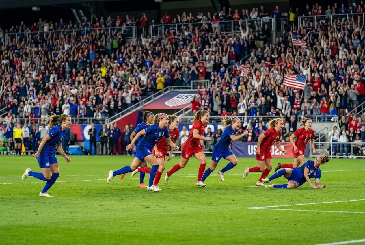 Women soccer players in action on a green pitch under evening floodlights with a packed, colorful crowd behind