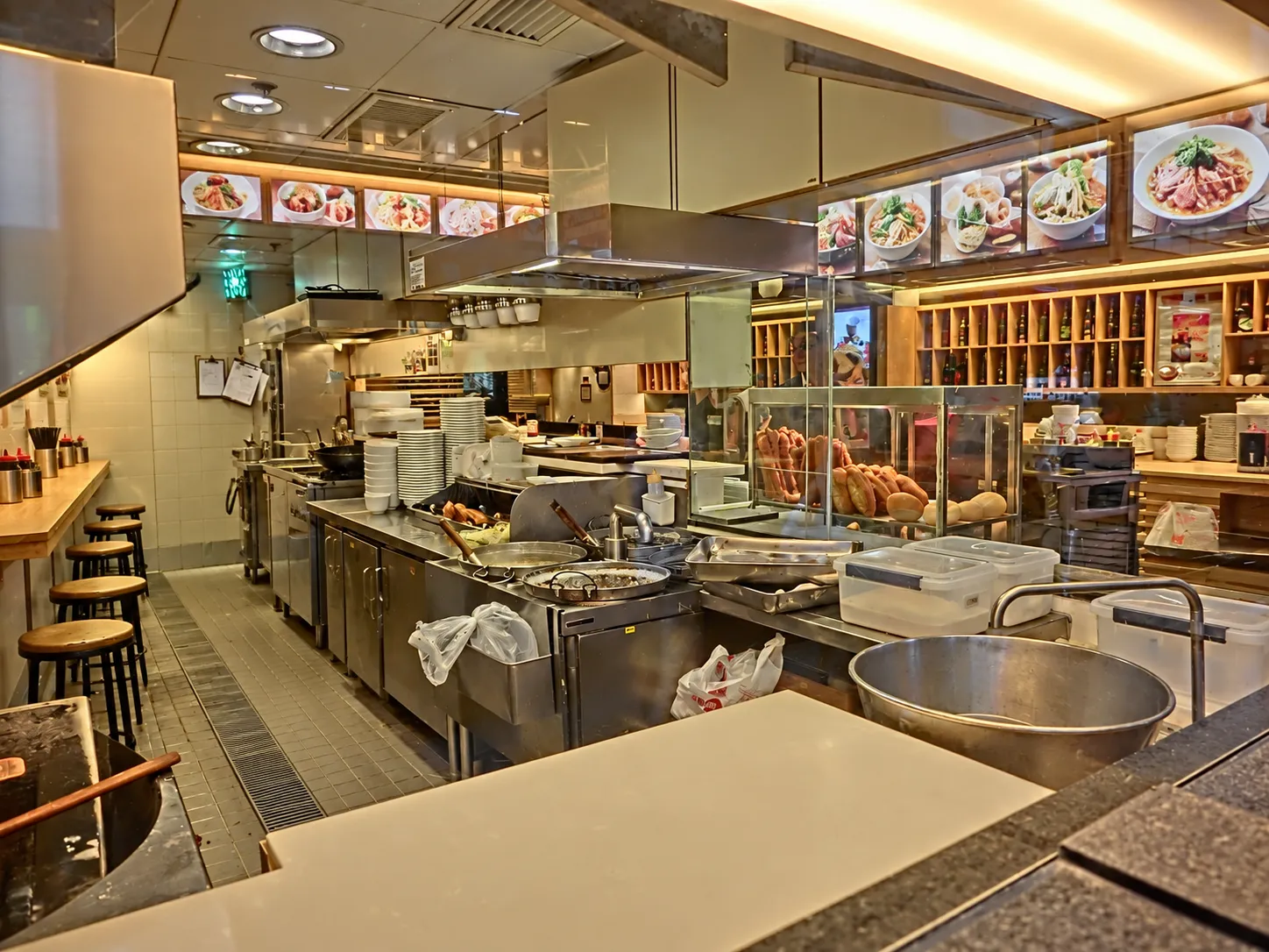 Narrow fast-casual noodle counter with register at the front, illuminated photo menu board above, stainless pass-through to the kitchen and empty stools along a wall shelf